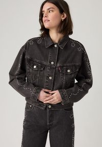 Woman wearing black denim jacket and jeans decorated with small silver studs, looking to the side with hands clasped at waist.