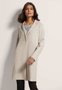 Einen cremefarbenen Mantel aus strukturiertem Stoff mit langen Ärmeln und Reverskragen. Über einem hellgrauen, mit Pailletten verzierten Oberteil getragen.