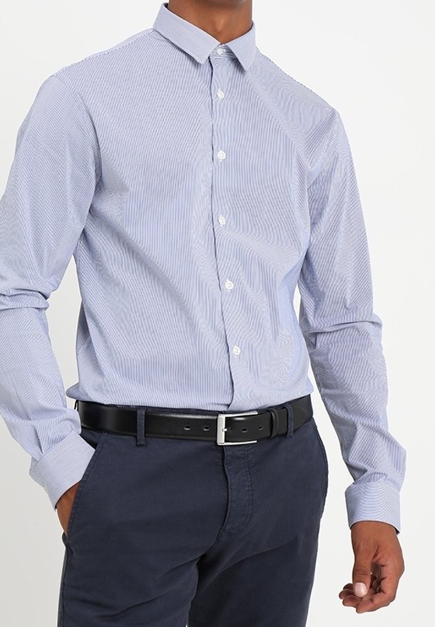 Homme portant une chemise à rayures bleu clair rentrée dans un pantalon bleu foncé avec une ceinture noire, debout avec une main dans la poche.