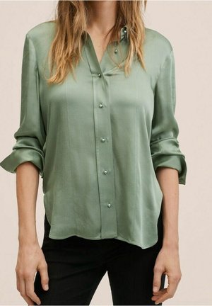 Vrouw draagt een losse, lichtgroene satijnen blouse met parelmoerknopen en opgerolde mouwen, gecombineerd met een zwarte broek.