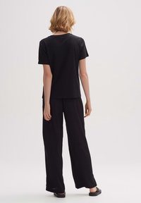 Svart bomullströja och vida byxor, båda i avslappnad passform, visas på en modell sedd bakifrån, med platta skor.