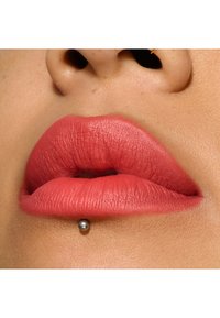 Matte koral læbestift på fyldige læber med synlig tekstur; en lille grå læbepiercing placeret under underlæben. Sløret hudbaggrund.