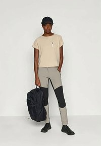Beige T-Shirt mit kurzen Ärmeln und Brusttasche, graue und schwarze, enganliegende Hose sowie einen schwarzen Rucksack, kombiniert mit einer schwarzen Kappe und Schuhen.