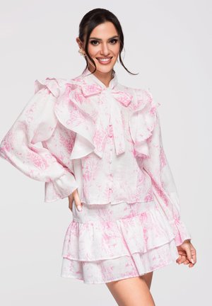 Femme souriante en robe blanche à manches longues avec imprimé floral rose, épaules à volants, nœud au col et jupe à étages, posant sur un fond uni.