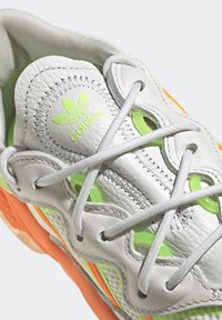 Valkoinen nahkainen tennari, jossa vihreitä yksityiskohtia, teksturoituja päällysteitä, hopeanväriset nauhat ja kirkkaan oranssi yksityiskohta pohjassa. Kengässä on Adidas-logo.