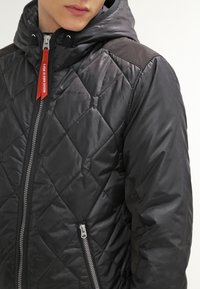 Svart vadderad jacka med huva, med ett diagonalt mönster, framdragkedja och en kontrasterande röd detalj på dragkedjans drag.