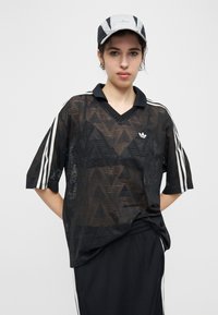 Νεαρό άτομο που φοράει μαύρο διάφανο πουκάμισο Adidas με λευκές ρίγες, μαύρο παντελόνι που ταιριάζει και μαύρο και άσπρο καπέλο Adidas, ποζάροντας με τα χέρια πίσω από την πλάτη.