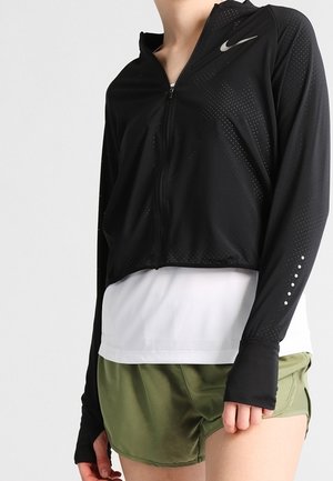 Zwarte cropped zip-up jas van lichtgewicht stof, met een zilveren Nike-logo en een geperforeerd patroon. G gedragen over een witte top en groene shorts.