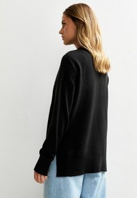 Czarny sweter dzianinowy o luźnym kroju, z długimi rękawami i ściągaczami. Tył ma nieco dłuższy krój i gładką teksturę.