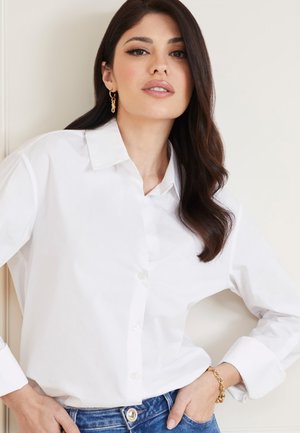 Camicia bianca con colletto, texture liscia, abbinata a jeans in denim blu e un braccialetto dorato. Capelli acconciati in onde morbide.