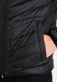 Svart pufferjacka med dragkedjestängning, diagonalt quiltat mönster och sidoficka. Materialet verkar lätt och glatt.