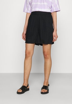 Nainen, jolla on mustat laskostetut shortsit, mustat ristikkäiset sandaalit ja violetti-valkoinen batikkikuvioinen lyhythihainen paita, seisomassa harmaalla lattialla.