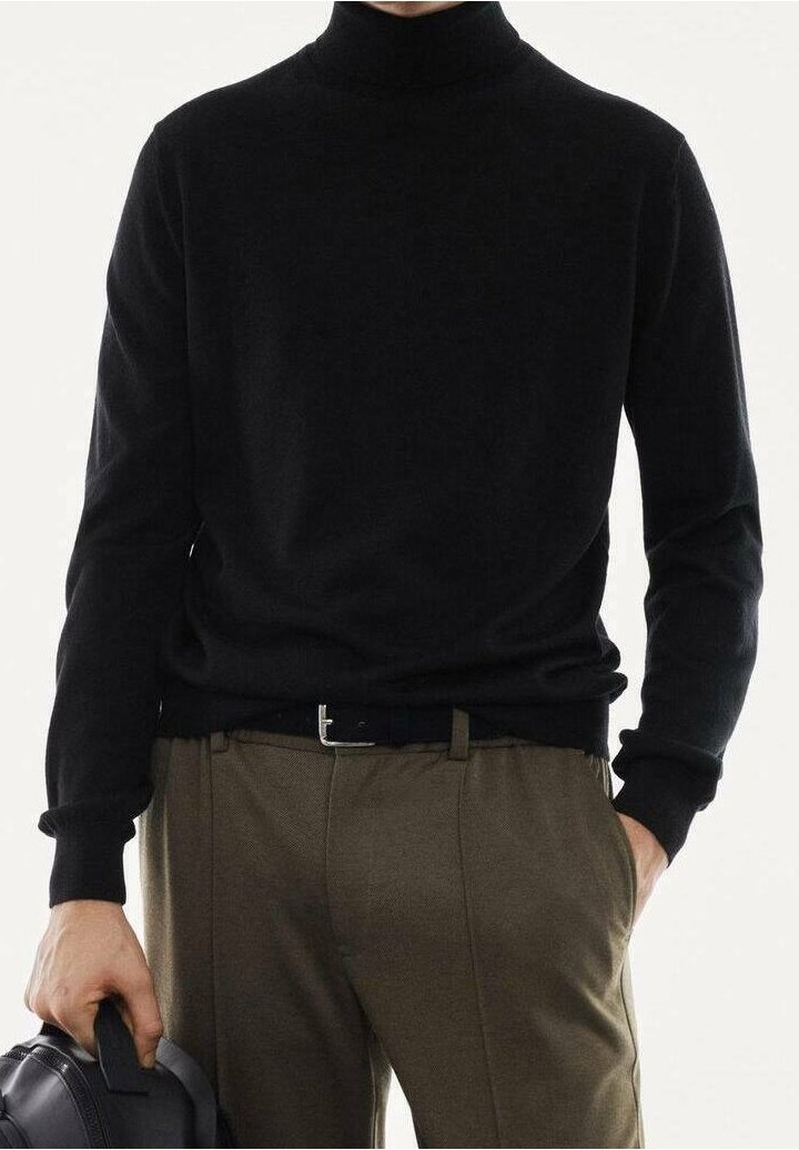 Personne portant un col roulé noir et un pantalon marron, tenant un sac noir dans la main gauche, la main droite dans la poche.