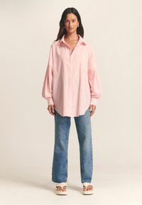 Light pink striped button-up shirt with a chest pocket, loose fit, long sleeves, paired with blue straight-leg jeans and cream sandals.