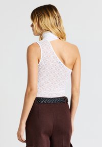 Donna con capelli biondi indossa un top in pizzo bianco a spalla sola e pantaloni marroni a vita alta con una cintura intrecciata nera, di spalle.