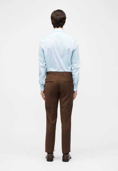Άνδρας που φορά ανοιχτό μπλε πουκάμισο, καφέ tailored παντελόνια και σκούρα καφέ επίσημα παπούτσια, στέκεται με την πλάτη στραμμένη σε ένα απλό λευκό φόντο.
