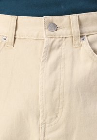 Beige cotton trousers with a zipper fly, metal button, and stitched detailing. Features front pockets and a textured weave pattern.