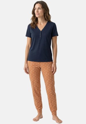 Vrouw staat blootsvoets, draagt een marineblauw henley T-shirt met korte mouwen en oranje joggerbroek met bloemmotief op een effen achtergrond.