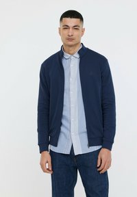 Veste zippée navy en tissu lisse, dotée d'un col montant, de poignets côtelés et d'un logo subtil. Portée sur une chemise à rayures bleues.