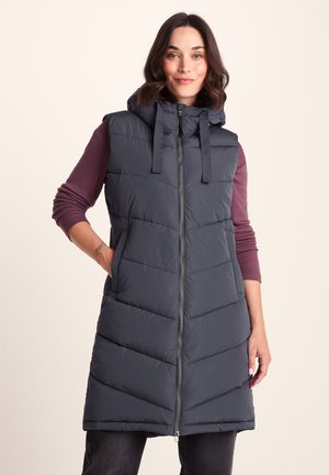 Woman wearing long sleeveless dark blue padded vest with hood over a mauve long-sleeve top, standing with one hand in pocket.