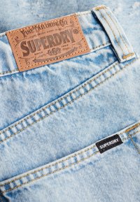 Jean en denim bleu clair en gros plan montrant des coutures cousues, une boucle de ceinture, une étiquette en cuir de la marque Superdry et une petite étiquette en tissu noir Superdry sur la poche.