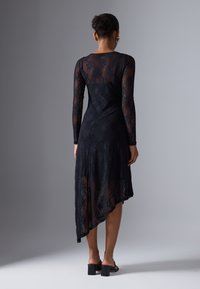 Robe en dentelle noire à manches longues, présentant un ourlet asymétrique et une texture transparente. Motif floral détaillé sur l'ensemble du tissu.