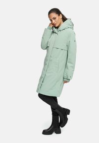 Mintgroene waterdichte parka met een capuchon, losse pasvorm, zijzakken en een middellange snit, gecombineerd met zwarte enkellaarzen.