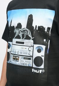 Melna T-kreklu ar grafiku, kurā attēlota pilsētas siluets ar sudraba pantheru, kas atrodas uz vintage audio sistēmas, ar zilu graduējošu fonu.