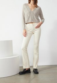 Hellgraue, kurz geschnittene Strickjacke mit V-Ausschnitt und Knopfleiste, kombiniert mit cremefarbenen, ausgestellten Jeans und schwarzen Schuhen mit glänzender Oberfläche.