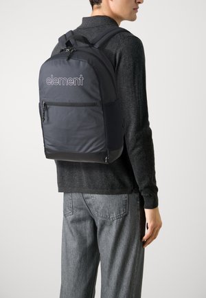 Mochila de nylon negra con logo, que cuenta con un bolsillo frontal con cremallera, correas acolchadas y acentos de malla, llevada en el hombro por una persona vestida con un atuendo gris.