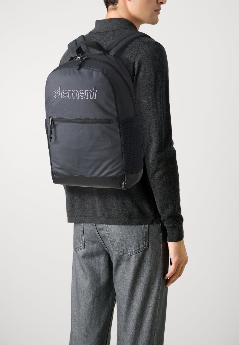 Zaino nero in nylon con logo, dotato di tasca frontale con zip, spallacci imbottiti e dettagli in rete, indossato a spalla da una persona in outfit grigio.