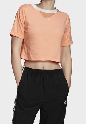 Cropped peach t-shirt with a white collar, featuring a small white adidas logo centered on the chest. Short sleeves with rolled cuffs.