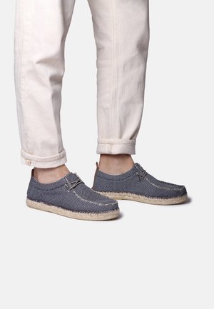 Zapatos de tela azul marino con un tejido texturizado, que cuentan con una suela de esparto beige y cordones mínimos, combinados con pantalones claros enrollados.