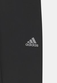 Pantalons de sport noirs avec une texture lisse. Présente un logo Adidas gris sur la cuisse gauche. Design simple, coupe ajustée.