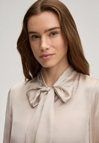 Beige satijnen blouse met een strik aan de kraag. Gladde textuur en gestructureerde pasvorm. Licht weerkaatst op de stof, wat een subtiele glans creëert.