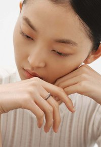 Anillo de plata con una banda delgada que presenta una superficie texturizada, llevado en una mano con dedos largos. Tonos de piel suaves y acabado natural en las uñas.