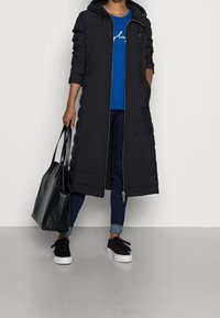 Sort polstret lang frakke med hætte, lynlås foran og quiltet tekstur. Kombineret med en blå skjorte, mørke jeans og sorte sneakers.