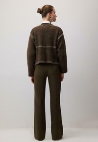 Femeie cu părul în coc poartă un pulover călduros de culoare maro cu modele albe și pantaloni evazați de un verde măsliniu închis, stând în fața unui zid gri simplu.