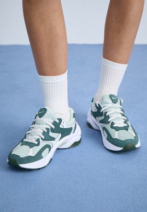 Persona con calcetines blancos hasta el tobillo y zapatillas Nike blancas con detalles verdes, de pie sobre una alfombra azul.