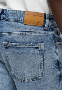 Τσέπη από ανοιχτό μπλε τζιν με μικρή ετικέτα σημαίας Tommy Jeans και καφέ δερμάτινο έμβλημα της μάρκας στη μέση της ζώνης, φορεμένη με λευκό τοπ.