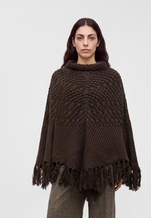 Femme aux longs cheveux foncés portant un poncho tricoté brun foncé texturé avec des franges sur un pantalon gris à carreaux, debout devant un fond uni.