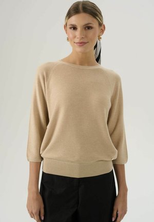 Beige Strickpullover mit Raglanärmeln, gerippten Bündchen und Saum. Weicher Stoff, lässige Passform und dezentes Muster. Getragen mit schwarzen Hosen.