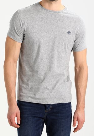 Mężczyzna noszący gładki jasnoszary t-shirt z krótkim rękawem, z małym ciemnym logo po lewej stronie klatki piersiowej oraz ciemnoniebieskie dżinsy.
