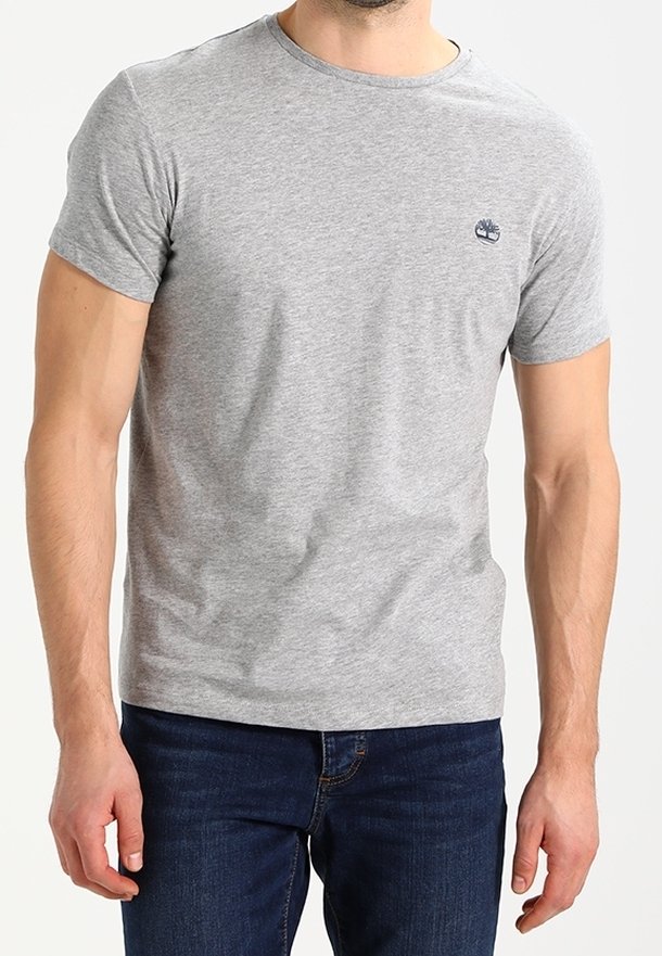 Homme portant un t-shirt uni gris clair à manches courtes avec un petit logo sombre sur le côté gauche de la poitrine et un jean bleu foncé.