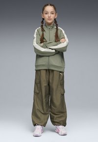 Veste olive à fermeture éclair avec des rayures blanches sur les épaules, associée à un pantalon cargo olive ample. Baskets rose clair avec un design texturé.