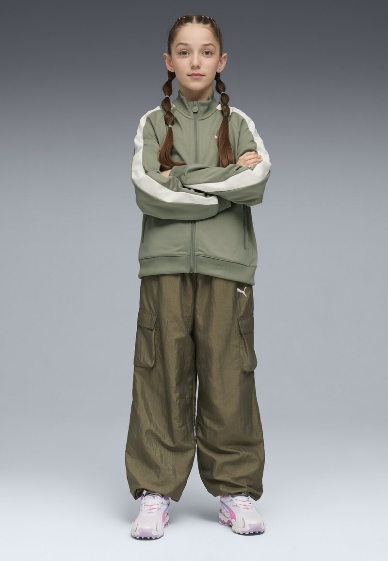Veste olive à fermeture éclair avec des rayures blanches sur les épaules, associée à un pantalon cargo olive ample. Baskets rose clair avec un design texturé.