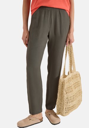 Person iført olivengrønne bukser, beige slip-on sko, der holder en stor beige vævet taske, med en orange top delvist synlig.