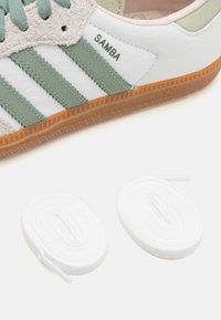 Vita lädersneaker med gröna och grå accenter, med tre gröna ränder, texturerad tunga och två vita snören i närheten.