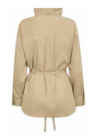 Chemise beige à manches longues avec col mandarin, plis au dos et taille nouée. Confectionnée en tissu léger, elle possède des poignets boutonnés pour la fermeture.