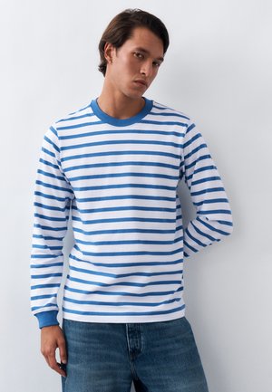 Νεαρός άνδρας με σκούρα μαλλιά, φορώντας μπλε και λευκή μπλούζα με οριζόντιες ρίγες μακριάς μανίκας και μπλε τζιν, πάνω σε απλό λευκό φόντο.